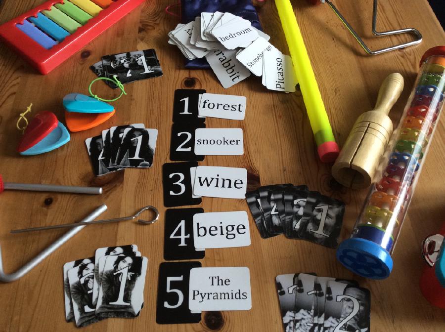A photograph of a boardgame I made as part of my PhD. It's called 'art of noises'. Photo shows several cards laid out with words on like 'forest', 'snooker', 'wine', and is surrounded by some colourful plastic instruments
