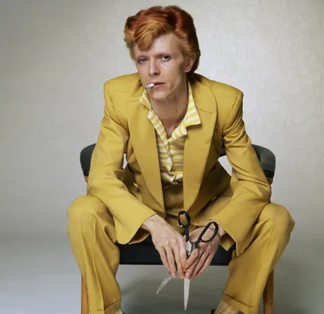 David Bowie in a mustard coloured suit, sitting on a chair, holding some scissors