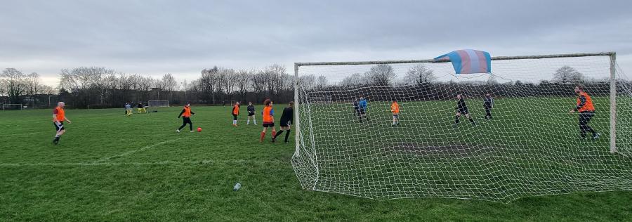 A photo of a football field on an overcast day with people kicking a ball around. There is a trans flag draped over the goalmouth